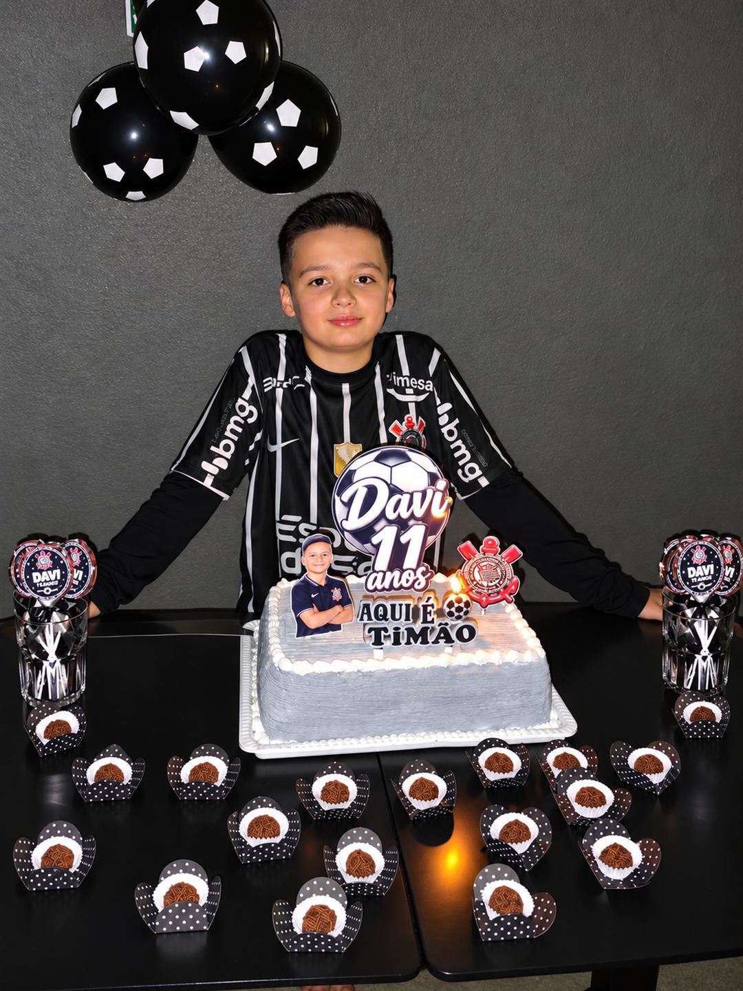 Aniversariante de 11 anos com camisa do Corinthians atrás do bolo temático, com docinhos na mesa e balões de futebol