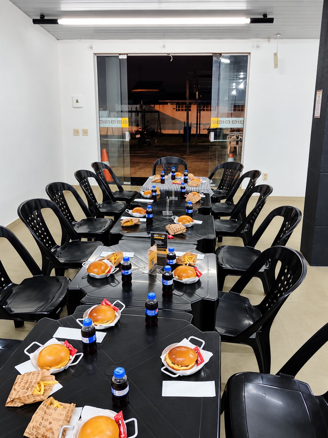 Salão da DIO arrumado para a festa, com hambúrguer, batata e refrigerante em cada lugar das mesas