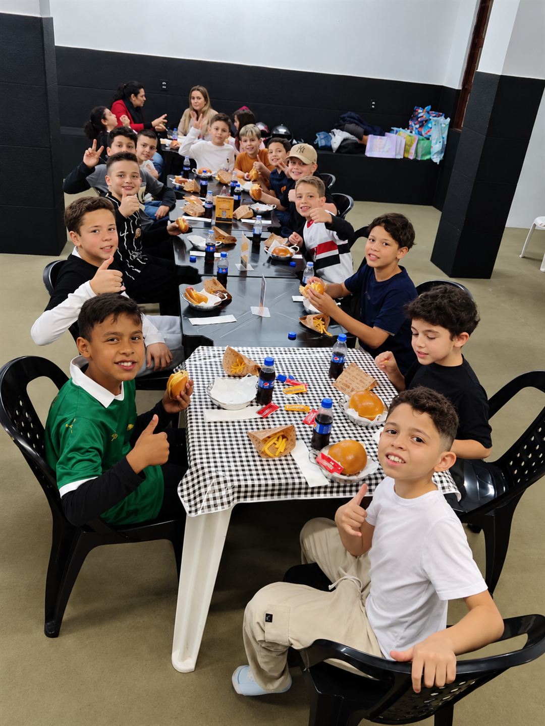 Crianças em uma mesa comprida fazendo joinha enquanto comem hambúrguer e batata