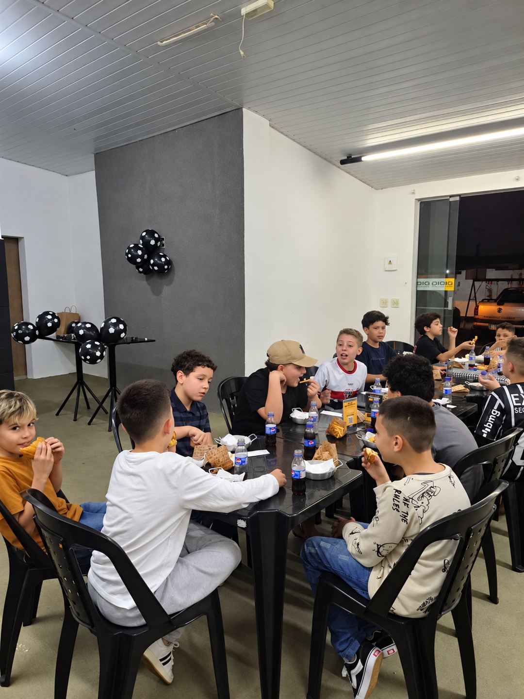 Crianças conversando e comendo em volta das mesas do salão, com balões de futebol ao fundo